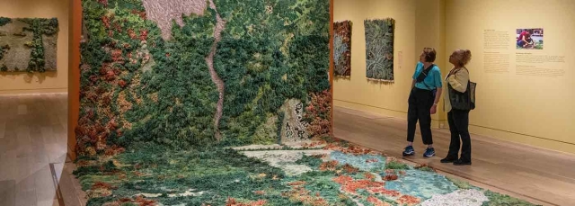 Two adults look at a large green and brown carpet artwork that drapes onto the floor.