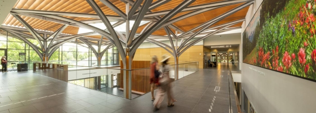 Top floor open atrium of the Freyer - Newman Center with windows on the left and a big LCD screen on the right. Blurred images of people that are walking along the pathway. 