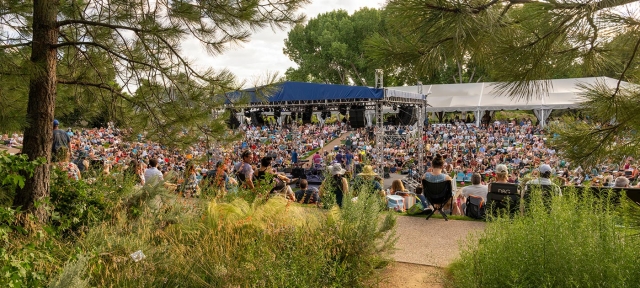 Tents at an outdoor summer concert with many people sitting on the grass looking at the stage.